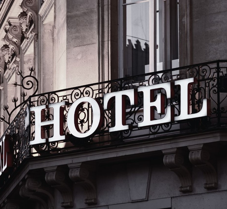  A classic hotel facade with ornate wrought iron balcony railings. Large white letters spelling “HOTEL” are mounted on the balcony, contrasting against the stone building behind.