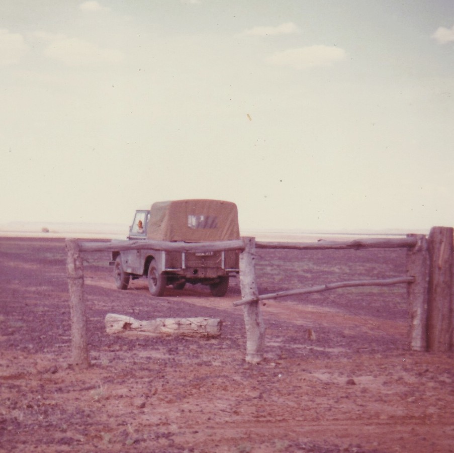 Old Land Rover with a canvas cover parked on a dirt track beyond a rough timber gate, in a flat, dry outback landscape under a pale sky.