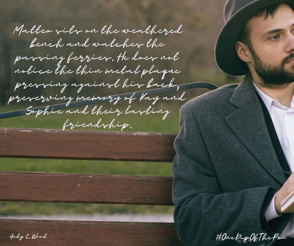 Alt text: A contemplative image shows a bearded man in a dark coat and hat seated on a weathered wooden bench beside a river or waterfront, looking toward unseen passing ferries. Overlaid handwritten text reads: “Matteo sits on the weathered bench and watches the passing ferries. He does not notice the thin metal plaque pressing against his back, preserving memory of Kay and Sophie and their lasting friendship.” The image is softly lit and reflective in tone, with the hashtags “#OneDipOfThePen” and the name “Andy C. Wood” included in the corners.